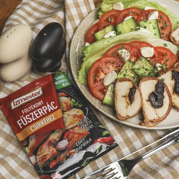 Aszalt szilvával göngyölt csirkemell felszeletelve, salátával tányéron felülnézetből. Mellette látható egy tasak folyékony fűszerpác csirkéhez.