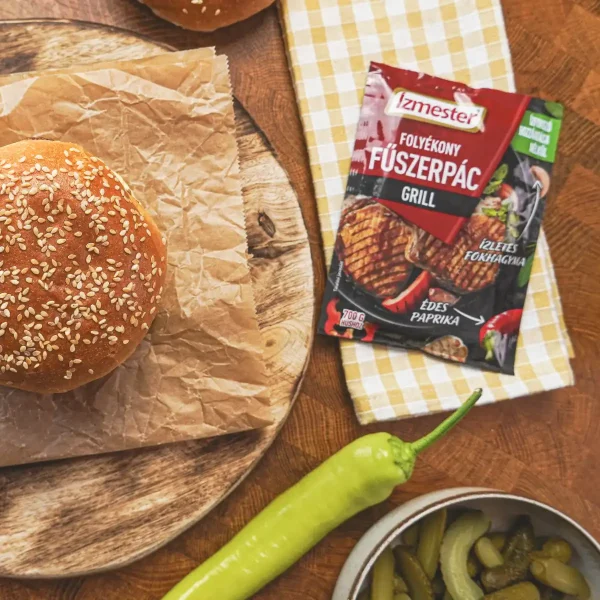 Szezámmagos buciban elkészített sertésburger papíron és fadeszkán tálalva konyhaasztalon. Mellette az Ízmester grill folyékony fűszerpác tasakja látható felülnézetből.