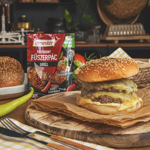 Szaftos sertésburger papíron és fadeszkán tálalva konyhaasztalon. Mellette az Ízmester grill folyékony fűszerpác tasakja látható.