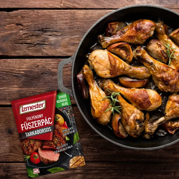 Sült csirkecombok felülnézetből fekete vasserpenyőben és mellette egy tasak Ízmester tarkaborsos folyékony fűszerpác tasak