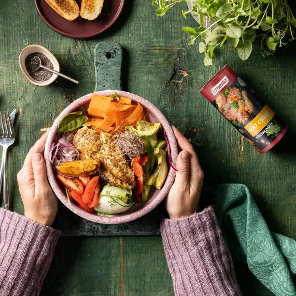 Egészben sült csirkemell salátaágyon rózsaszín tányérban, amit egy női kéz fog körbe, konyhaasztalon, mellette sült csirke szórófejes fűszerkeverékkel és zöldfűszerrel