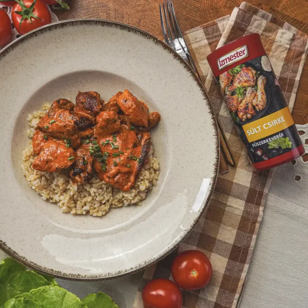 Paradicsomos paprikás csirke barna rizzsel bézs színű tányéron egy konyhaasztalon, mellette egy doboz sült csirke szórófejes fűszerkeverék