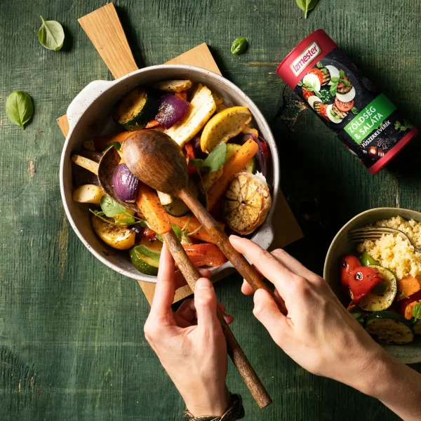 Sült zöldségek egy tálban, sült lilahagyma, fokhagyma, krumpli, cukkini és kaliforniai paprika, mellette saláta fűszerkeverék, egy kéz a salátából vesz ki