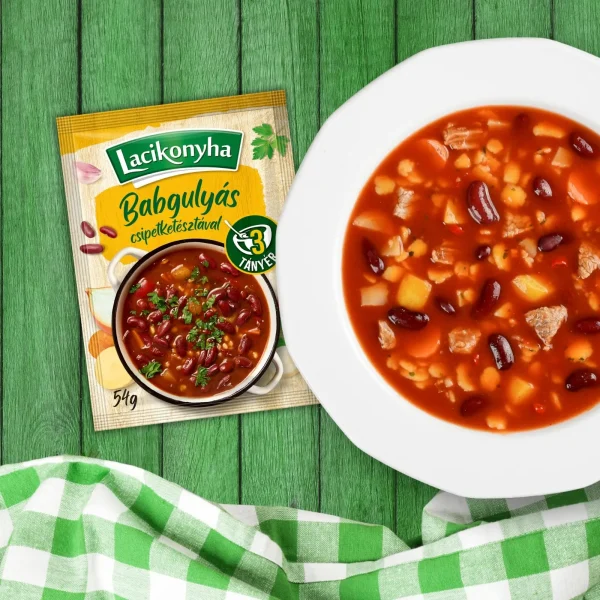 Babgulyás csipetkével házias stílusú levespor tasak faerezetes asztalon, mellette fehér tányérban babgulyás és zöld-fehér kockás terítővel