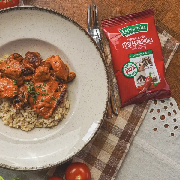 Paprikás-paradicsomos csirkemell falatok barna rizs fehér tányéron mellette egy tasak csemege édes magyar fűszerpaprika