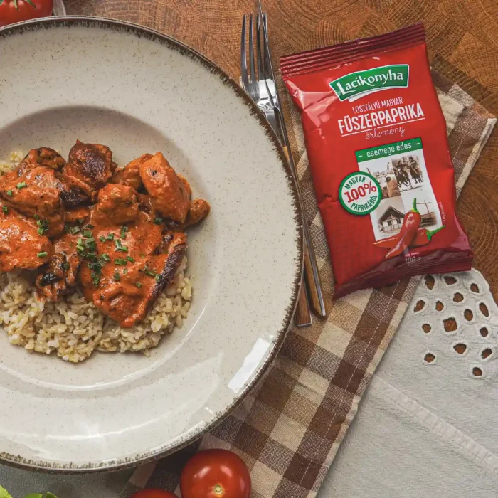 Paprikás-paradicsomos csirkemell falatok barna rizs fehér tányéron mellette egy tasak csemege édes magyar fűszerpaprika