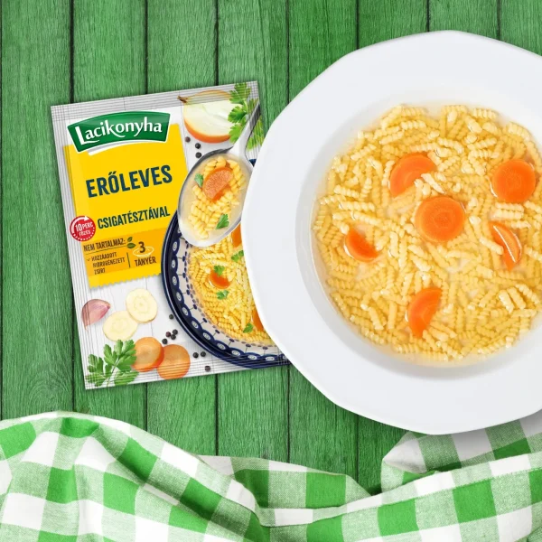 Erőleves csigatésztával házias stílusú levespor tasak faerezetes asztalon, mellette fehér tányérban erőleves és zöld-fehér kockás terítővel