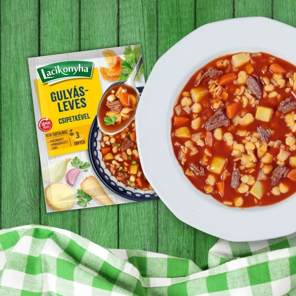 Gulyásleves csipetkével házias stílusú levespor tasak faerezetes asztalon, mellette fehér tányérban gulyásleves és zöld-fehér kockás terítővel