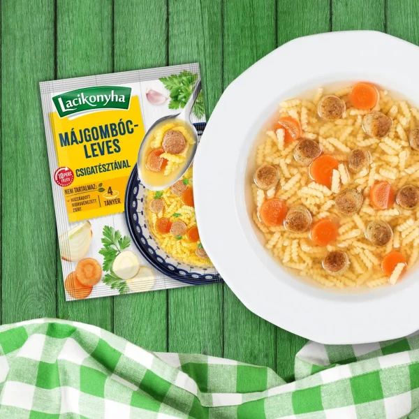 Májgombócleves házias stílusú levespor tasak faerezetes asztalon, mellette fehér tányérban májgombócleves és zöld-fehér kockás terítővel