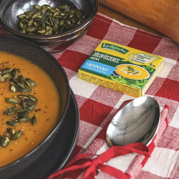 sütőtökkrémleves tökmaggal sötét tányérban, kanállal mellette egy doboz zöldfűszeres kocka