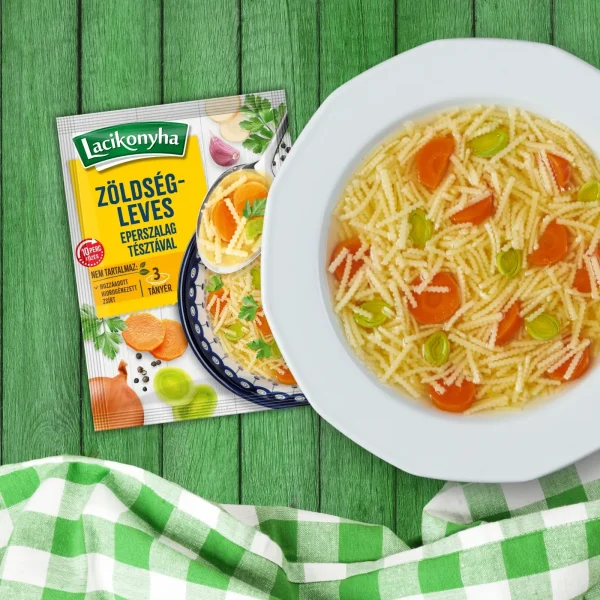 zöldségleves eperszalag tésztával házias stílusú levespor tasak faerezetes asztalon, mellette fehér tányérban zöldségleves és zöld-fehér kockás terítővel