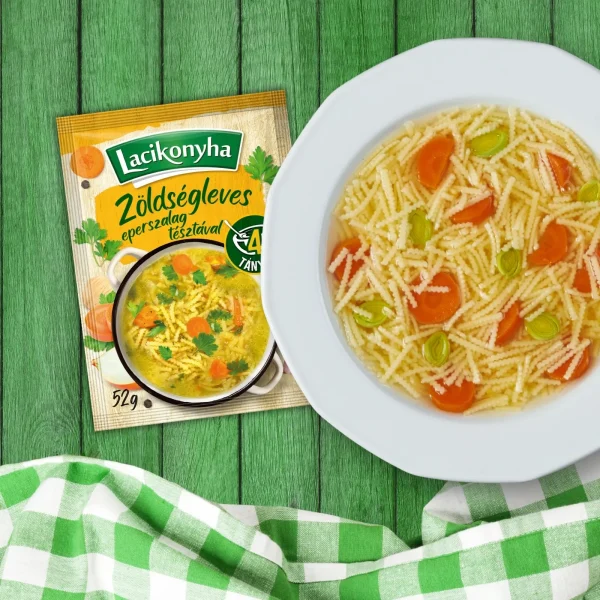 zöldségleves eperszalag tésztával házias stílusú levespor tasak faerezetes asztalon, mellette fehér tányérban zöldségleves és zöld-fehér kockás terítővel