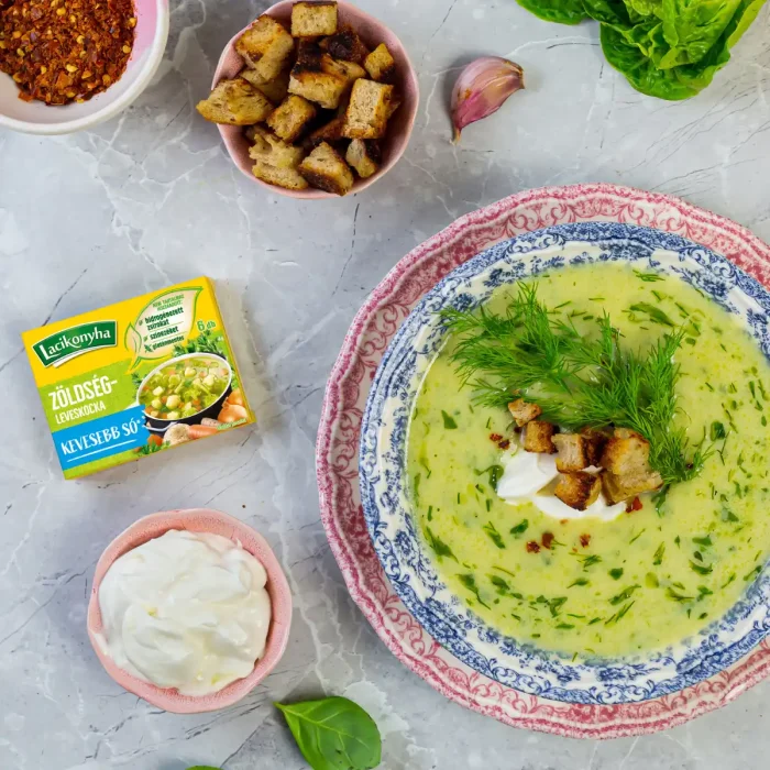 Cukkini krémleves tányérban mellette kis tálnyi tejföl és egy doboz zöldfűszeres kocka