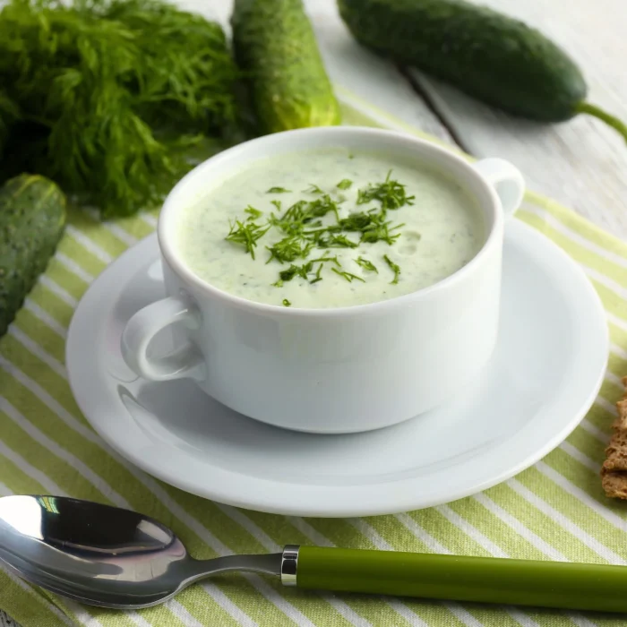 Hideg uborkaleves fehér leveses tányérban tálalva asztalterítőn, mellette dekorációként uborka, friss kapor, kanál