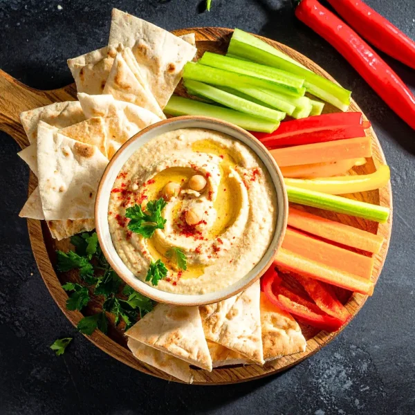 Krémes hummusz tál felülnézetből egy fa deszkán, mellette pita kenyér és zöldséghasábok