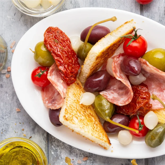 Előétel antipasti szárított paradicsommal, olívabogyóval, gyöngyhagymával, sonkával és pirított kenyérrel fehér tányéron tálalva