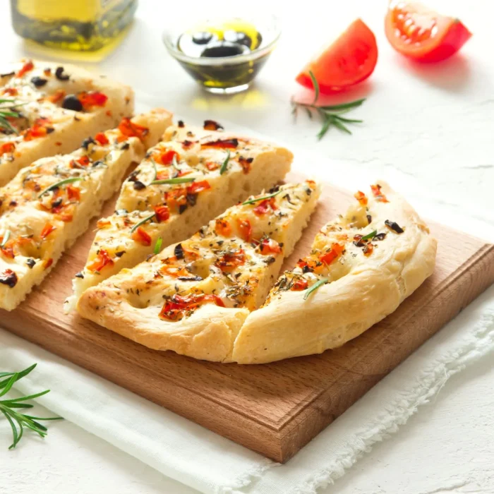 Focaccia házi kenyér fűszerekkel fa deszkán, paradicsomszeletekkel, rozmaringgal tálalva