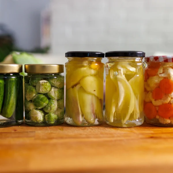 Egy fa asztalon befőttes üvegek sorakoznak, bennük savanyúságok: savanyú uborka, kelbimbó, paprika, karfiol, zöldség savanyúságok.