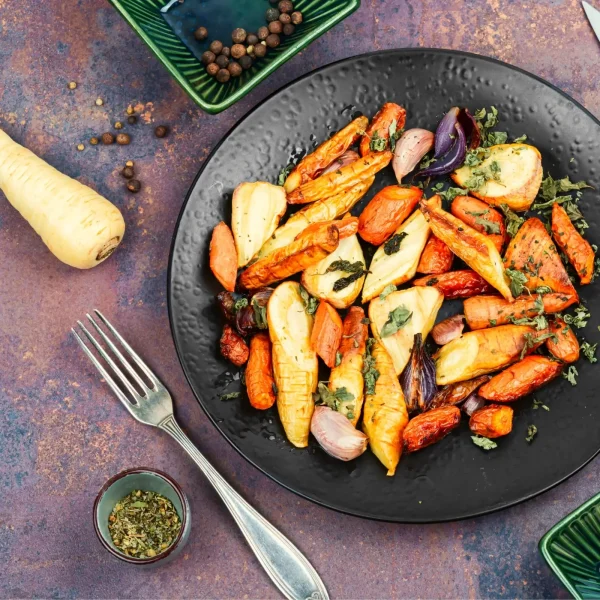 Vegyes sült gyökérzöldségek fekete tányérban felülnézetből, mellette villával